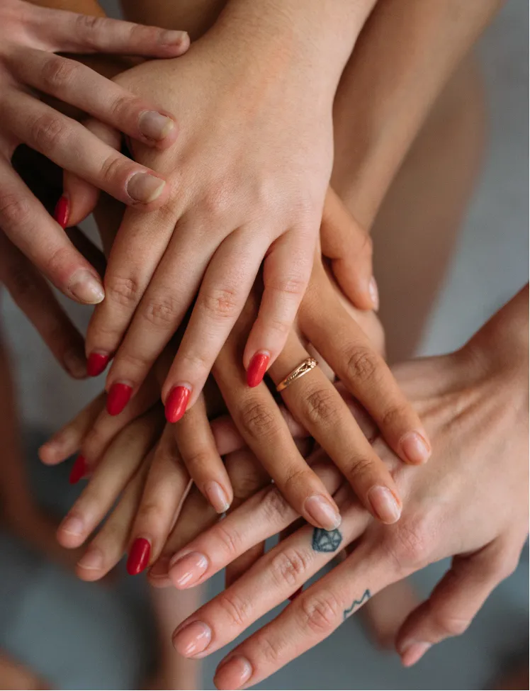 community-2 A group of diverse hands stacked together, some with nail polish and one hand with a ring and a tattoo.