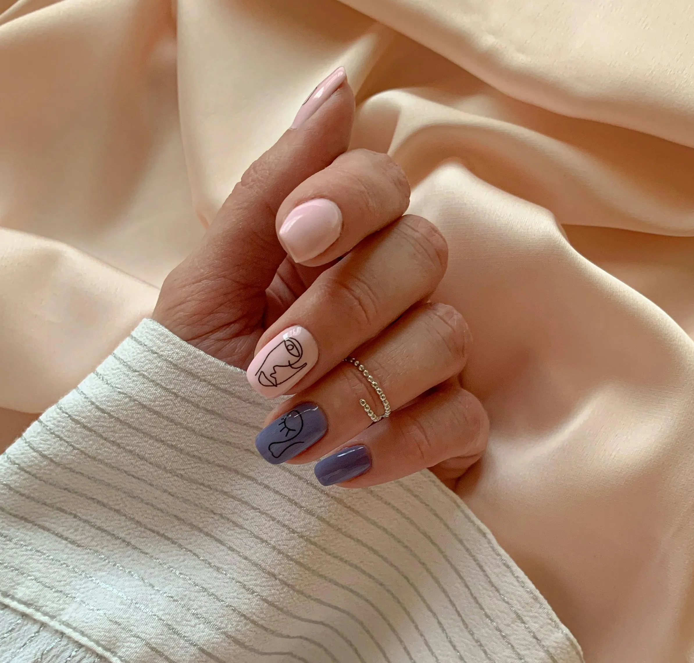 gel-x A hand with purple and pink nail art, two nails featuring minimalist line face designs, on a beige satin background.