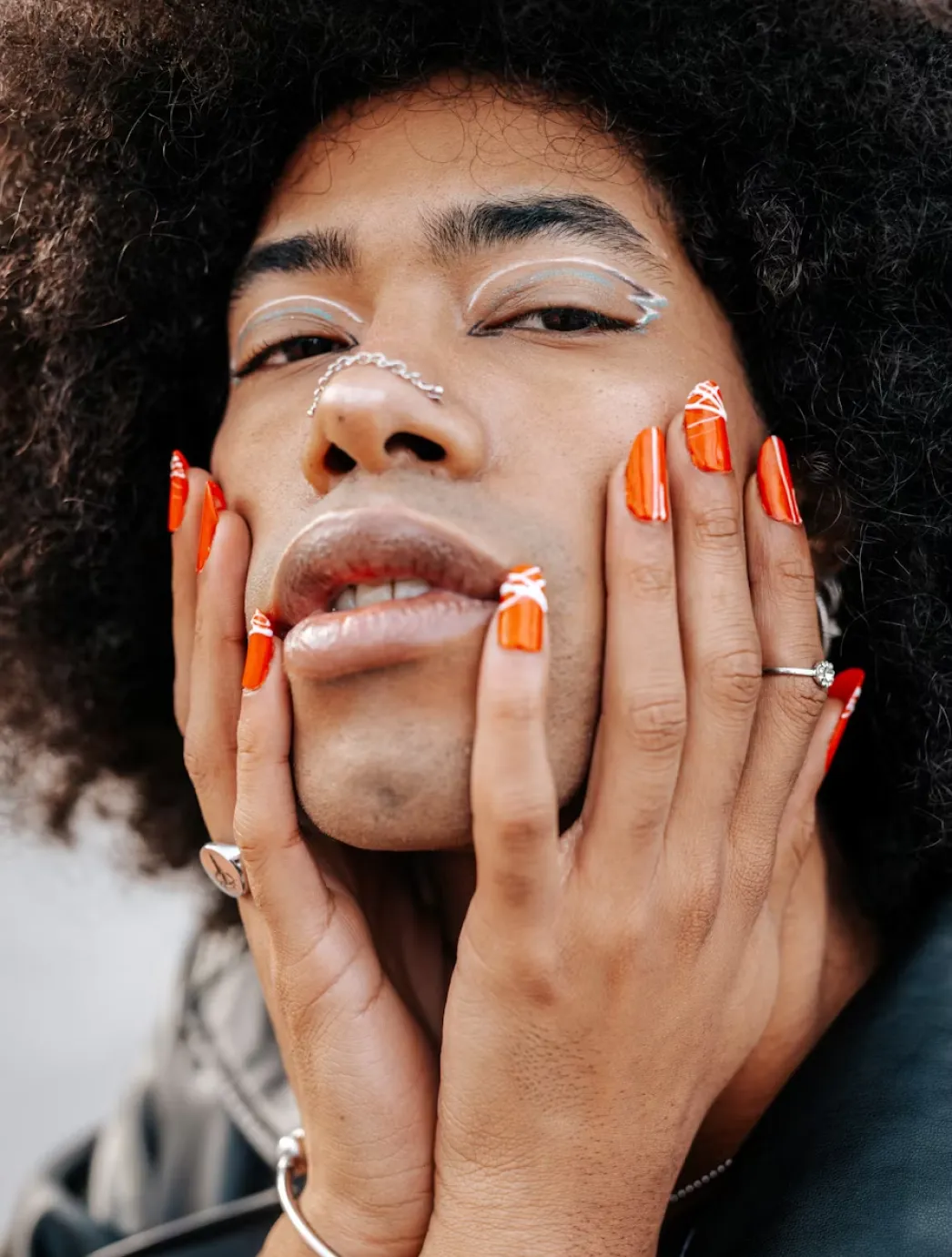 image 2 Person with bold orange nails and abstract white eyeliner touching their face, looking directly at the camera.
