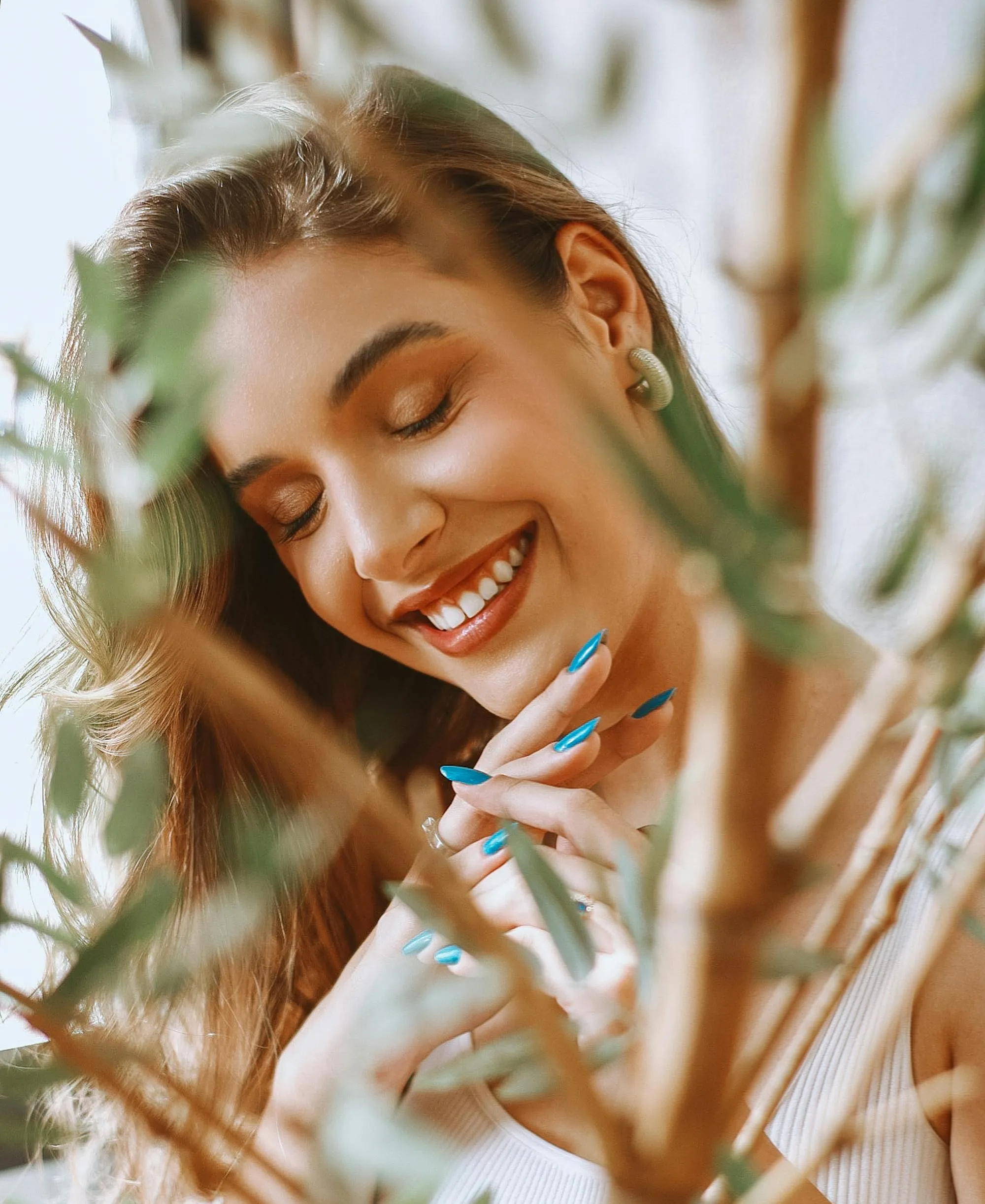 philo-1 Woman smiling with eyes closed, wearing blue nail polish, framed by blurred green leaves in the foreground.